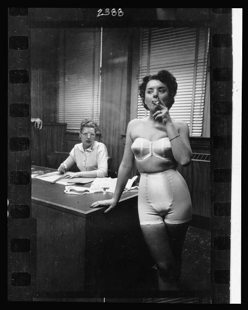 21.-Lingerie-model-wearing-a-girdle-and-strapless-bra-smoking-in-an-office-in-the-background-a-woman-sits-at-a-desk.jpg